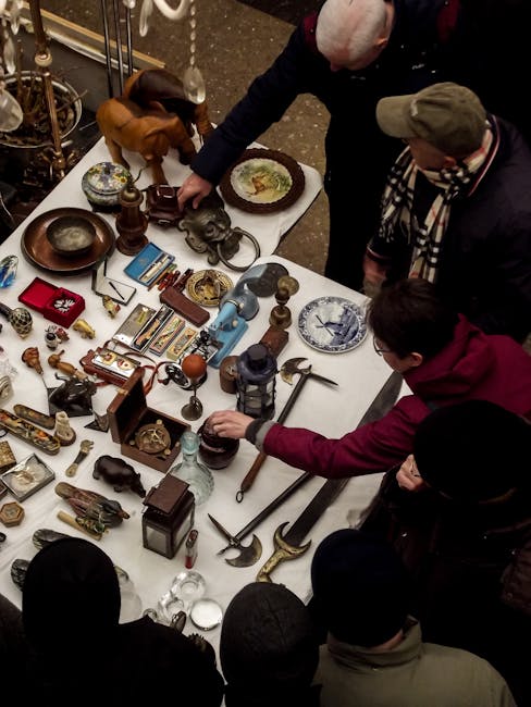 An overhead view of a group of five individuals gathered around a rectangular table covered with various antique and vintage items, including ornamental objects, small sculptures, clocks, framed photographs, and decorative plates. The individuals, dressed in casual winter clothing with some wearing hats and scarves, are actively inspecting and handling the objects, with one person reaching out to pick up an item. The scene appears to take place in an indoor setting, possibly at an antique fair or market, where collectors or buyers are evaluating potential purchases. The table is densely packed with unique items made from materials such as metal, wood, and glass, showing a range of textures from polished to aged surfaces. The environment includes a dark, speckled floor, and the background hints at a bustling, indoor marketplace or auction environment related to private disposal or on-site clearance, which aligns with independent rubbish removal or collection services offered by Rubbish Collection Waterloo.