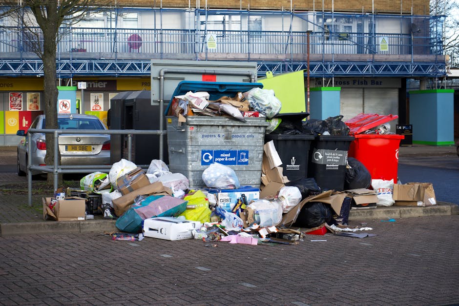 A collection of overflowing rubbish bins and gathered waste on a paved street area, including a large grey mixed cardboard and paper recycling container with its lid open, revealing assorted paper, cardboard boxes, and plastic bags spilling onto the ground. Surrounding the bin are numerous black, white, and red trash bags, some torn open, along with flattened cardboard boxes, plastic packaging, and loose paper scattered nearby. On the left side, a metallic grey car is parked close to a metal railing, with a small tree partially obscuring the scene. In the background, a building features a ground-floor storefront with signage and large windows, while an upper level is under construction or renovation, covered with blue scaffolding and protective netting. The area appears to be in an urban environment, with natural daylight illuminating the scene and highlighting the cluttered state of the refuse collection.