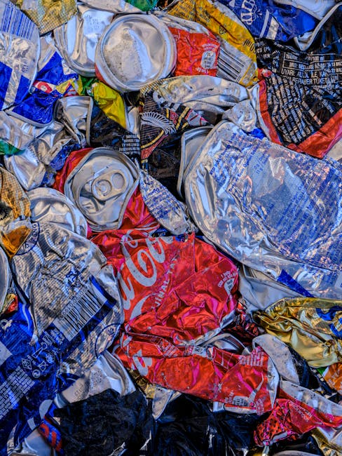 An overhead view of a group of five individuals gathered around a rectangular table covered with various antique and vintage items, including ornamental objects, small sculptures, clocks, framed photographs, and decorative plates. The individuals, dressed in casual winter clothing with some wearing hats and scarves, are actively inspecting and handling the objects, with one person reaching out to pick up an item. The scene appears to take place in an indoor setting, possibly at an antique fair or market, where collectors or buyers are evaluating potential purchases. The table is densely packed with unique items made from materials such as metal, wood, and glass, showing a range of textures from polished to aged surfaces. The environment includes a dark, speckled floor, and the background hints at a bustling, indoor marketplace or auction environment related to private disposal or on-site clearance, which aligns with independent rubbish removal or collection services offered by Rubbish Collection Waterloo.