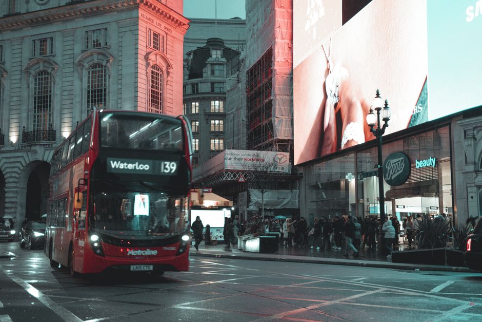 A busy urban scene at dusk showing a red double-decker bus on route 139 heading towards Waterloo, with reflective windows revealing the interior. The street is illuminated by streetlights, with some parked cars partially visible on the left side. In the background, tall historic and modern buildings feature large windows and detailed facades, with billboard advertisements on their upper levels. To the right, a glass-fronted shop, likely a retail or beauty outlet, displays a large digital advertisement showing a close-up of a person's legs in white clothing. A group of pedestrians, dressed for cool weather, is walking along the pavement in front of the shop, some waiting to cross or browsing. The environment suggests a central shopping and transit hub, where alternative waste handling and rubbish collection services like Rubbish Collection Waterloo may support local waste disposal needs amid the city's vibrant activity, with the scene emphasizing urban cleanliness and organized public transport.