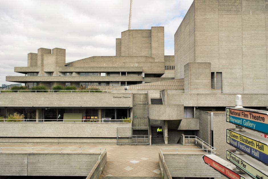 A large, multi-level concrete building with a modernist architectural style, featuring angular and block-like structures, expansive flat surfaces, and minimal decorative elements. The exterior surfaces have a rough, textured finish typical of concrete, with sections showing signs of weathering and slight discoloration. In the foreground, a paved open area includes a set of stairs and pathways leading to the building entrances, with a single worker wearing a high-visibility vest standing near the stairs, possibly involved in maintenance or clearance activities. To the right, a multi-colored directional signboard indicates nearby attractions such as the National Film Theatre, Hayward Gallery, Queen Elizabeth Hall, Royal Festival Hall, and Festival Pier, suggesting a cultural precinct. The scene is lit under an overcast sky, creating diffuse, natural lighting that emphasizes the building’s stark, utilitarian design. This environment appears to be part of a public or semi-public urban space, with the potential context of on-site waste management or clearance activities by independent waste removal services, consistent with the focus on rubbish and waste handling associated with large cultural venues.
