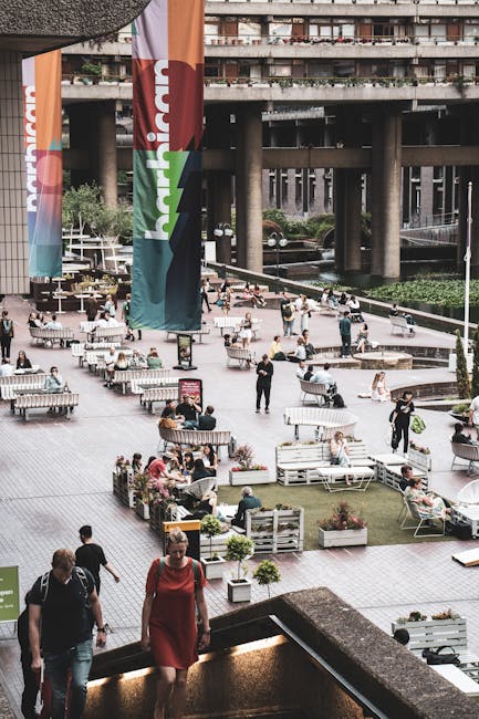 An open outdoor public space with a paved surface featuring several white metal and wooden benches, some of which have people sitting on them or walking nearby. The area includes potted plants in rectangular planters, arranged along the benches and walking paths. In the foreground, a woman in a red dress and a man with a backpack are ascending a step or staircase. The background showcases a large, multi-level concrete building with numerous balconies, and there are colorful vertical banners on poles that display the word 'heineken' in bold white and red letters. The scene is well-lit, suggesting daylight, and the environment appears lively with people engaging in leisure activities, which may relate to event setups or casual gatherings. The image gives an impression of a busy urban park or event space, with the potential for independent waste management or rubbish collection services like those offered by Rubbish Collection Waterloo, supporting the care of public areas and event cleanup efforts in urban sites similar to this setting.